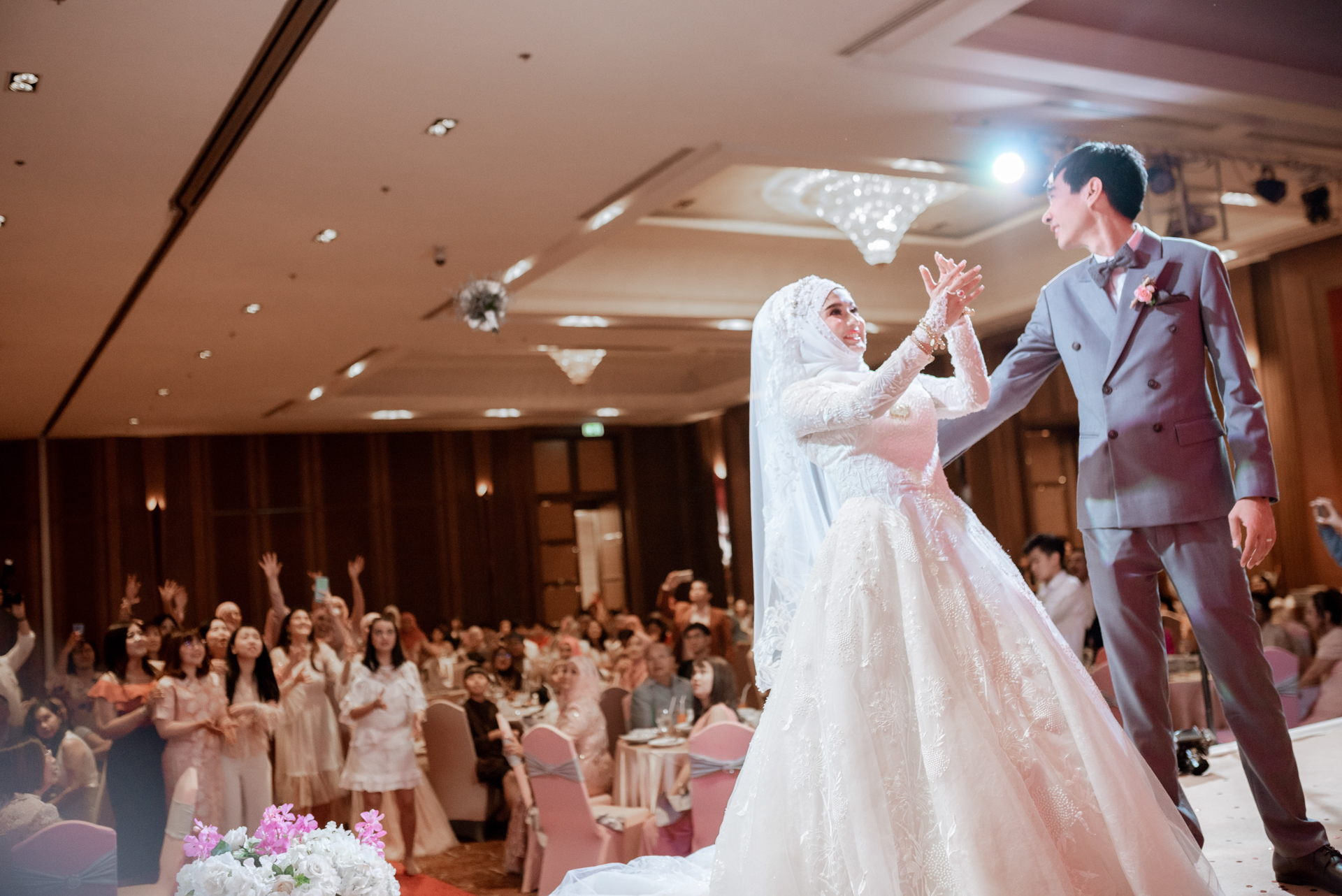 เจ้าบ่าวเจ้าสาวปรบมือด้วยกันบนเวที ท่ามกลางแขกจำนวนมากที่ปรบมือเชียร์และยกโทรศัพท์ถ่ายภาพ